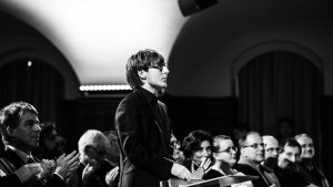 Chorleiter Cornelius Hofmann im Profil, zum Chor blickend, im Hintergrund Publikum