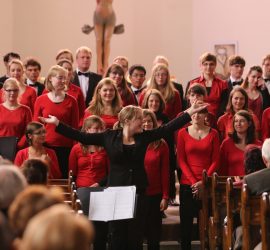 Chor in mehreren Reihen, davor Chorleiterin Ines Kaun zum Publikum gewendet, die Arme ausbreitend